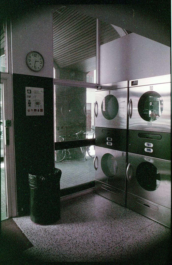why-choose-us A dimly lit indoor laundromat featuring multiple stainless steel washing machines and a visible clock.